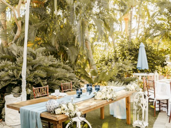 A rustic outdoor table setup with blue accents, white flowers, and lanterns in a tropical garden setting.