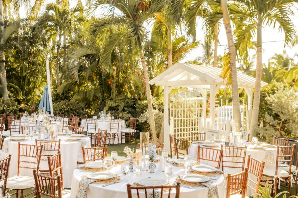 Outdoor wedding setup with round tables, white tablecloths, elegant decorations, and wooden chairs under palm trees in a garden setting.