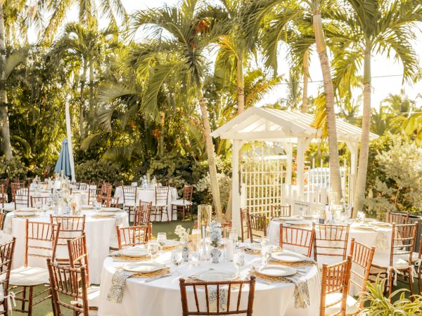 Outdoor wedding setup with round tables, white tablecloths, elegant decorations, and wooden chairs under palm trees in a garden setting.