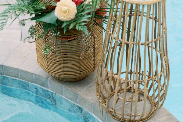 A colorful floral arrangement in a wicker basket is placed beside a wicker lantern on the edge of a pool.