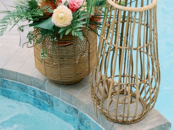 A colorful floral arrangement in a wicker basket is placed beside a wicker lantern on the edge of a pool.