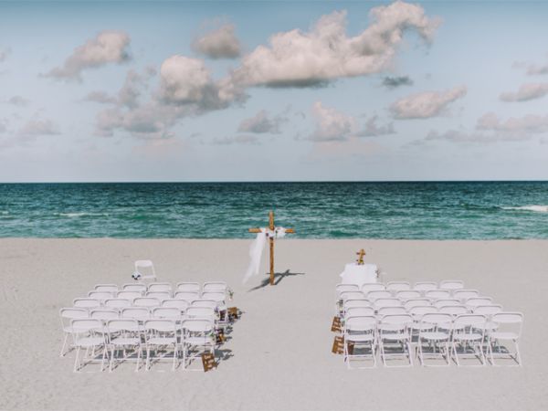 A beach wedding setup with white chairs and an arch facing the ocean under a partly cloudy sky.