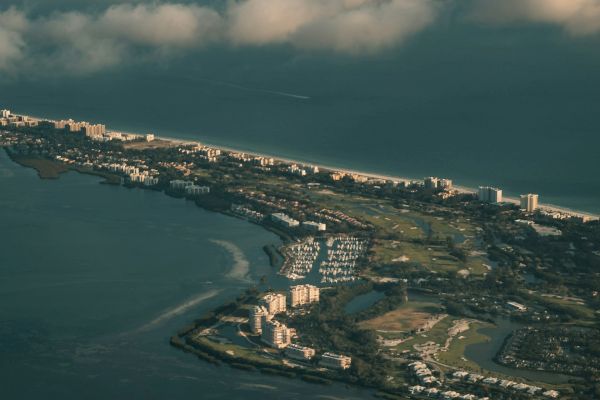 Aerial view of a coastal city with buildings, green spaces, and a marina, surrounded by the sea and partly cloudy skies.