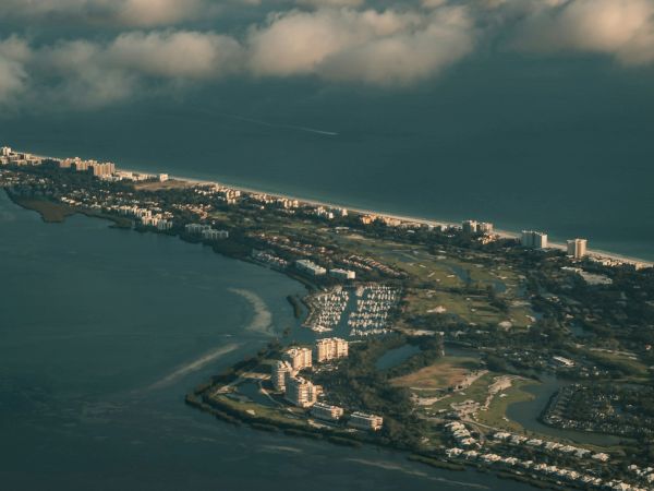 Aerial view of a coastal city with buildings, green spaces, and a marina, surrounded by the sea and partly cloudy skies.