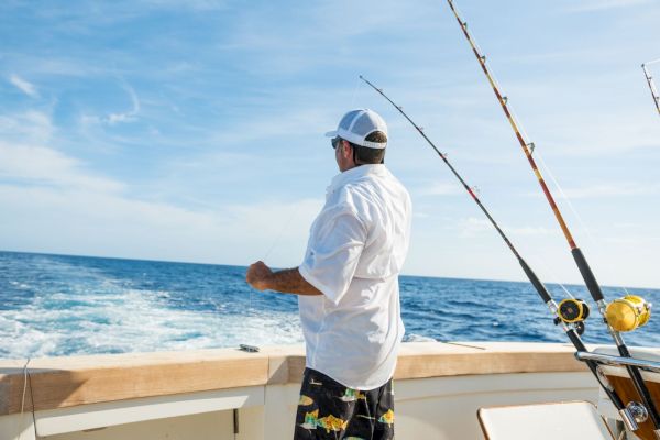 A person fishing at the back of a boat in the ocean, holding a line, with multiple fishing rods in view.