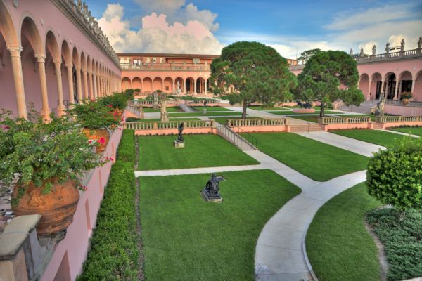 A courtyard with manicured lawns, sculptures, potted plants, and pink arched buildings in a classic architectural style.