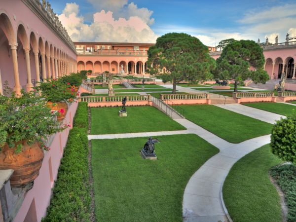 A courtyard with manicured lawns, sculptures, potted plants, and pink arched buildings in a classic architectural style.