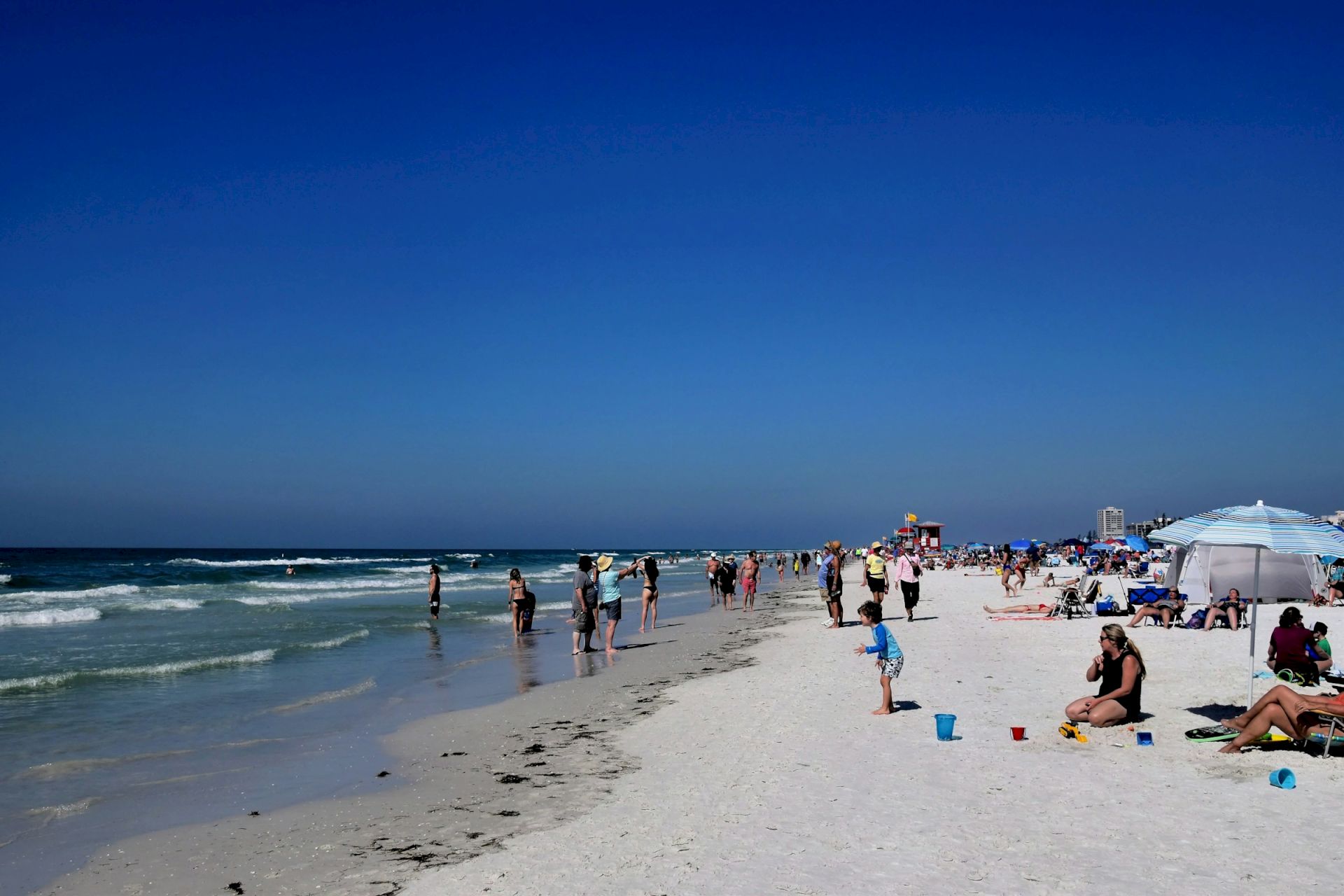 People enjoy a sunny day at the beach, with umbrellas and waves in the background, creating a lively and relaxing atmosphere.