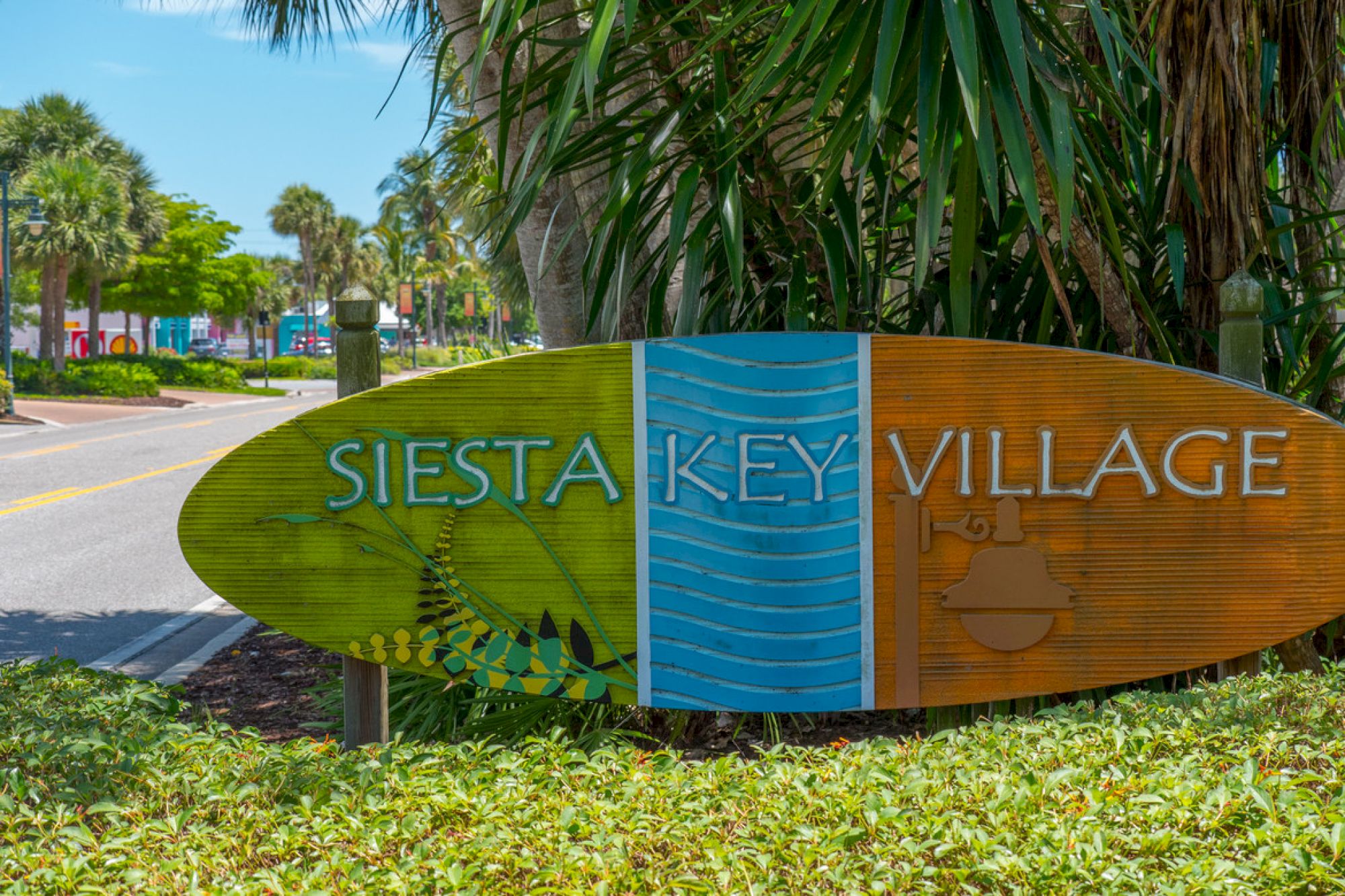 A sign for Siesta Key Village surrounded by greenery, with a road and palm trees visible in the background.