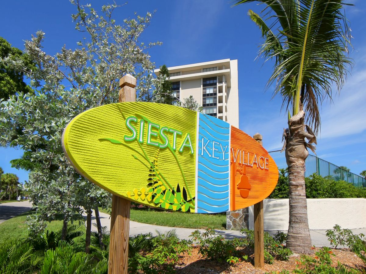 A colorful sign reads "Siesta Key Village" in front of trees and a modern building under a clear blue sky.