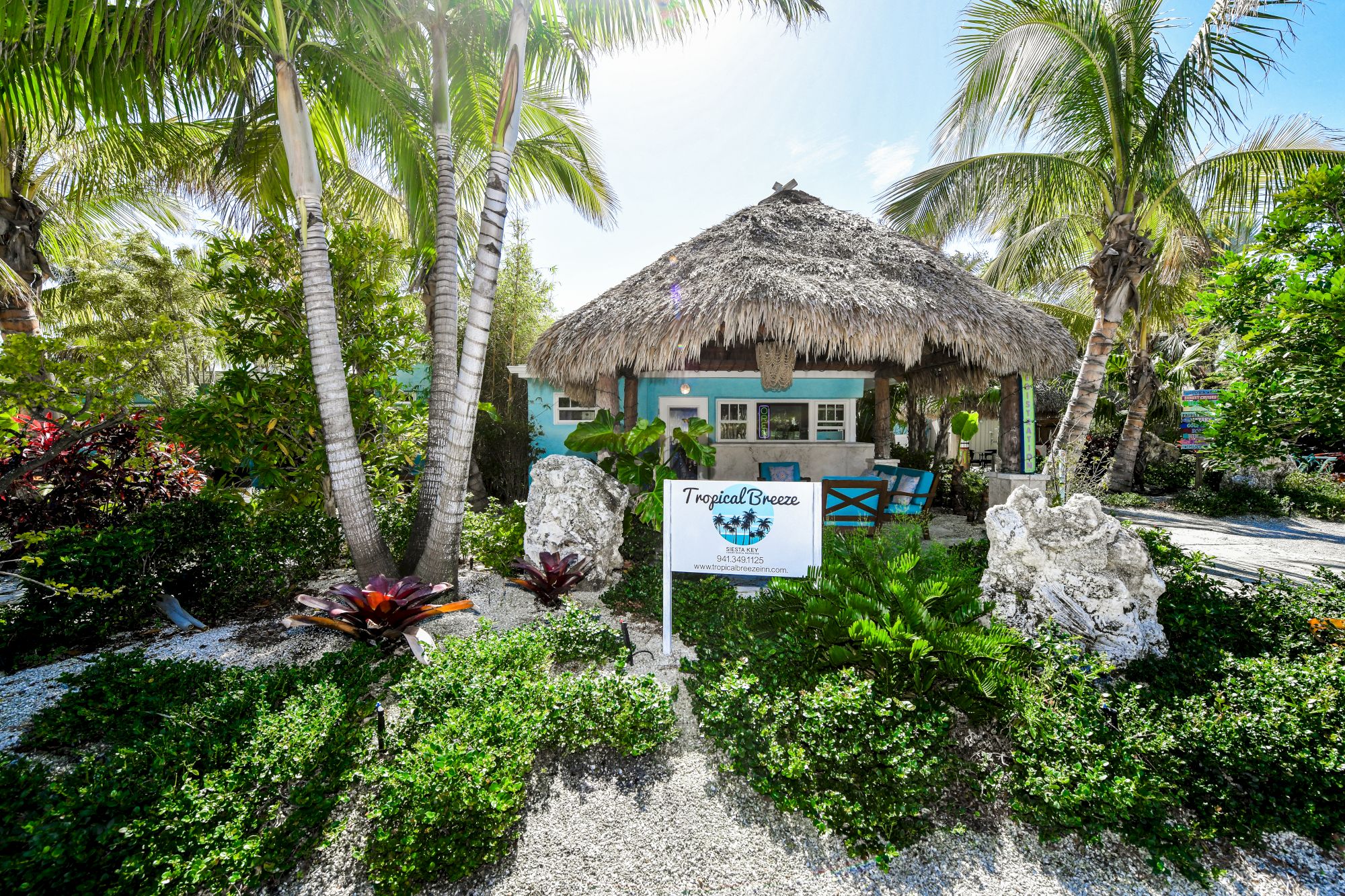 A tropical-themed building with a thatched roof surrounded by palm trees and plants; a sign stands in front with lush greenery around.