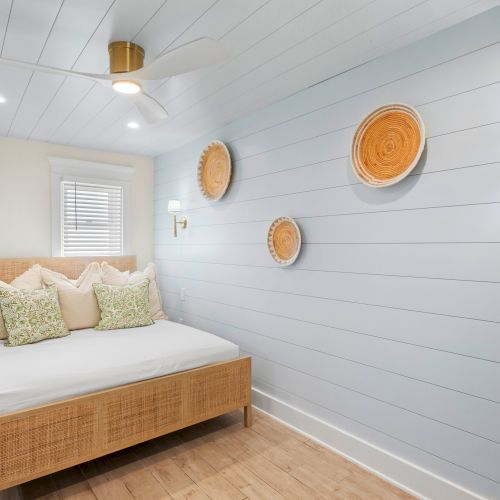 A cozy bedroom with a bed, decorative pillows, light blue paneled walls, wall sconces, and woven wall baskets for decoration.