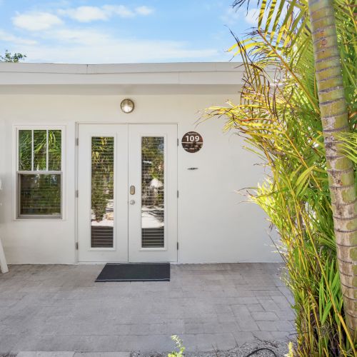 A white building entrance with double doors, palm plants, and a sign reading "109."