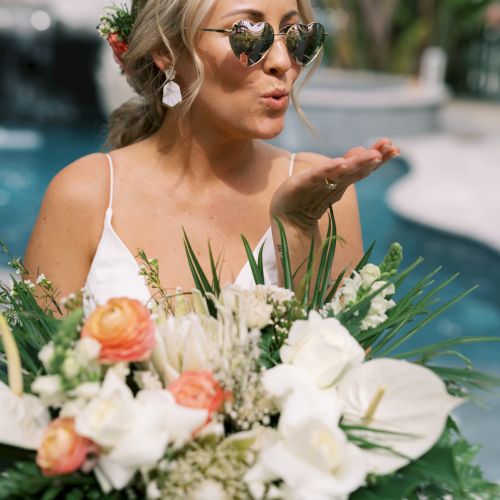 A woman in heart-shaped sunglasses holds a bouquet, blowing a kiss by the poolside.