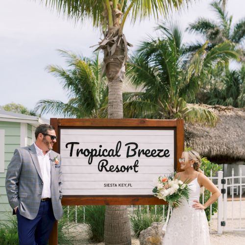 A couple stands beside a "Tropical Breeze Resort" sign in Siesta Key, FL, surrounded by palm trees.