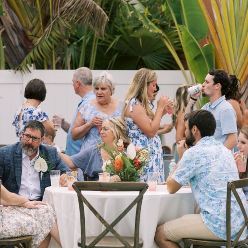 A group of people at an outdoor gathering, some seated around a table with a floral centerpiece, others standing and socializing, drinks in hand.