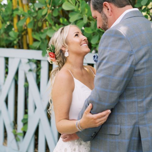 A couple is embracing and smiling at each other, surrounded by greenery, in a warm and affectionate setting.