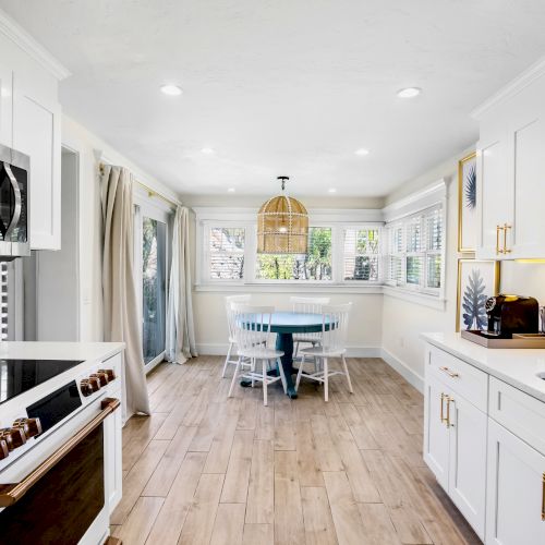A modern kitchen with white cabinets, wood floors, a round table, and large windows. There's a coffee maker on the counter.