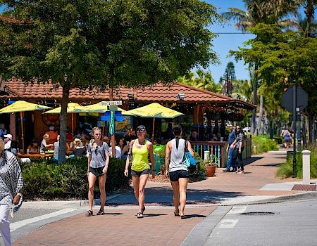 A busy street scene with people walking and dining outdoors under umbrellas on a sunny day, surrounded by trees and buildings.
