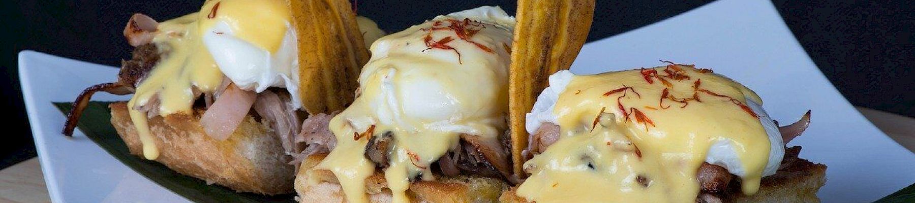 The image shows three pieces of bread topped with poached eggs, sauce, shredded meat, and garnished with plantain chips, served on a platter.