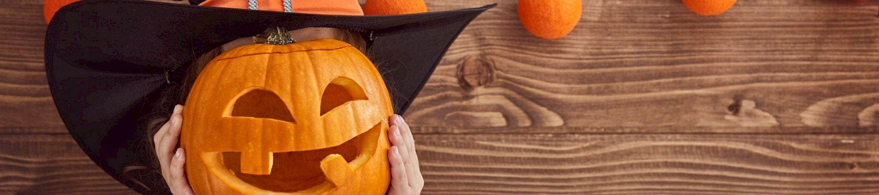 A person in a witch costume holding a carved jack-o'-lantern in front of their face, with orange decorations hanging on wooden background.