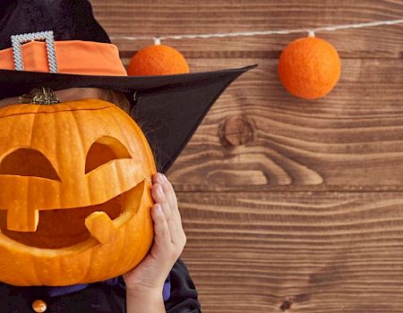 A person in a witch costume holding a carved jack-o'-lantern in front of their face, with orange decorations hanging on wooden background.