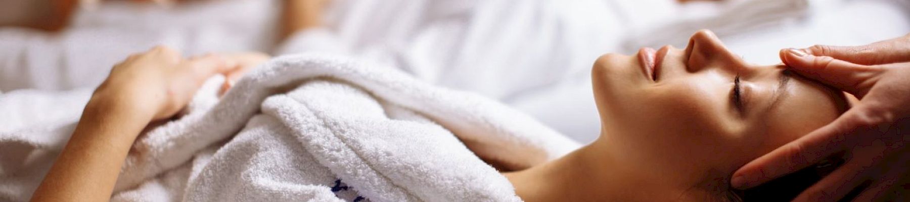 Two people in white robes are receiving face massages, lying on spa beds with relaxed expressions and gentle lighting.