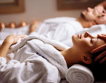 Two people in white robes are receiving face massages, lying on spa beds with relaxed expressions and gentle lighting.
