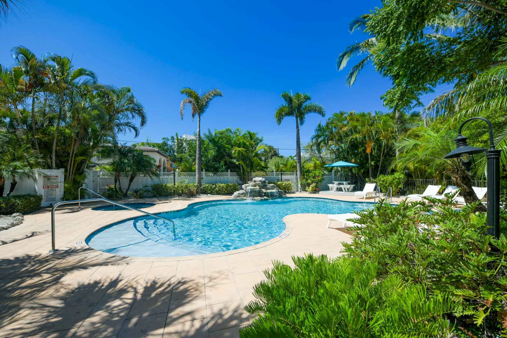 A swimming pool surrounded by lush greenery, palm trees, and lounge chairs under a clear blue sky in a tropical setting.
