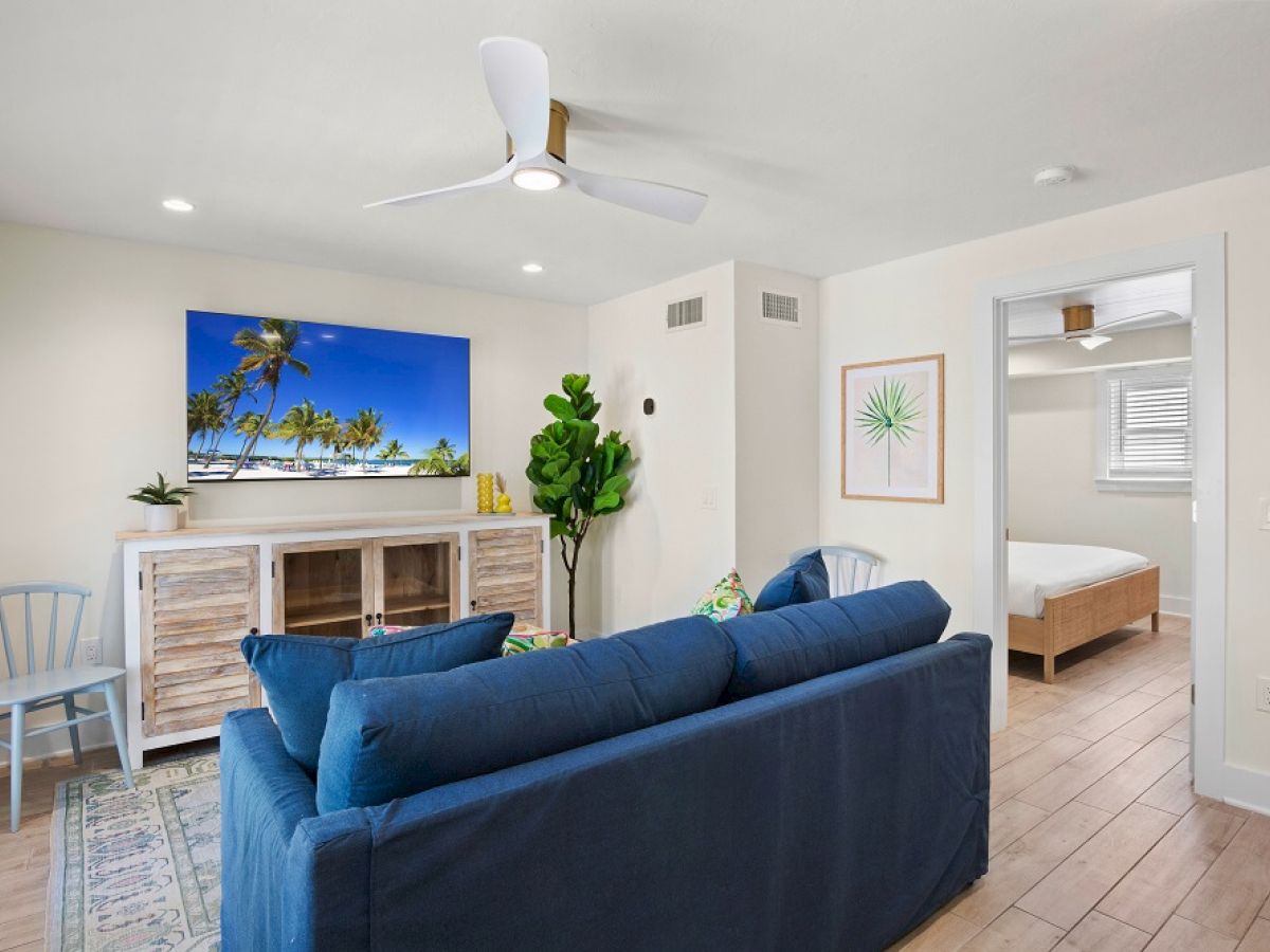A cozy living room with a blue sofa, TV, ceiling fan, and decor. A doorway leads to a bedroom with a bed.