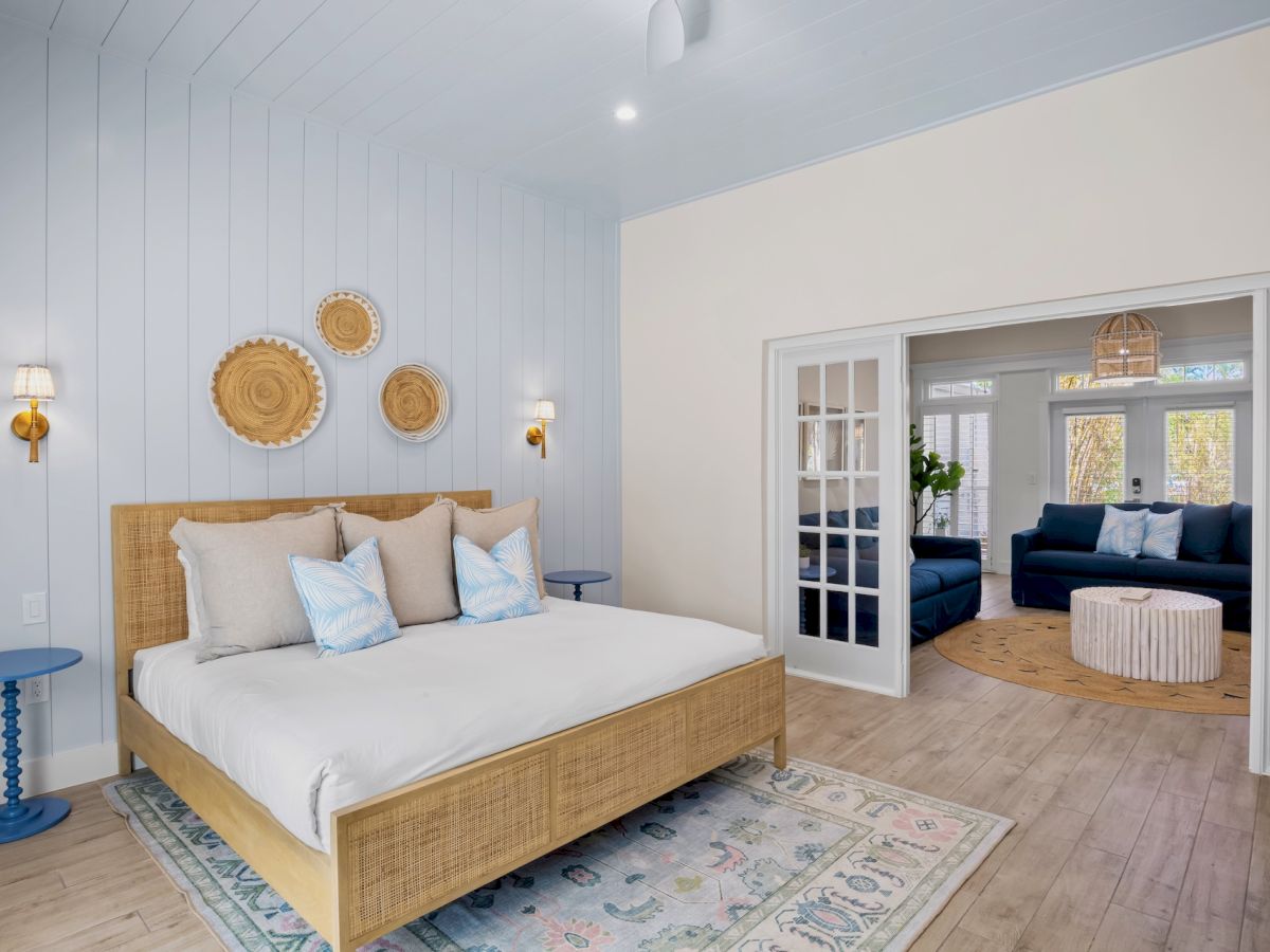 A bedroom with a bed, side tables, and wall decor; a sitting area is visible through French doors in a cozy, bright setting.